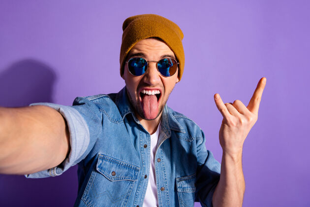 眼镜开朗疯狂摇滚迷的自拍照片 展示摇滚标志角状手指伸出舌头 穿着牛仔裤牛仔夹克 在紫色充满活力的背景下与世隔绝自拍时髦男人