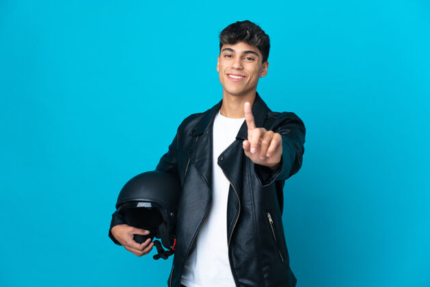 夹克一个戴着摩托车头盔的年轻人在孤立的蓝色背景上展示并举起一根手指男人头盔拇指