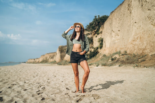 女性戴帽子的黑发女孩在沙滩上和岩石一起放松短裤户外快乐