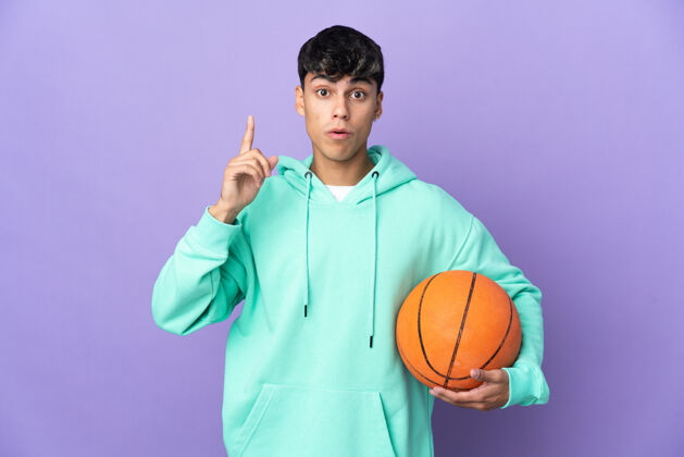 球一个年轻人在孤立的紫色背景上打篮球 想在举起手指的同时实现解决方案哇健康微笑