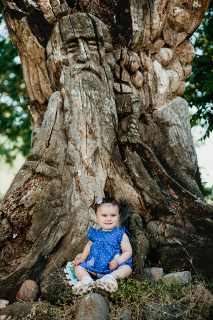 探索穿着蓝色裙子的时髦婴儿坐在老大树旁微笑着巨树乐趣休闲
