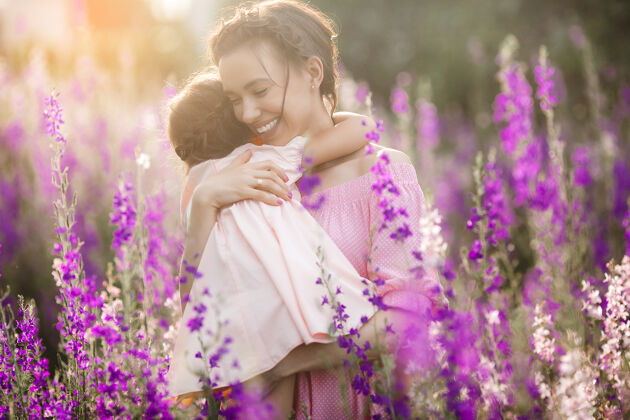 微笑年轻漂亮的妈妈和她的小女儿一起在户外玩耍花紫罗兰色花坛日落可爱母性