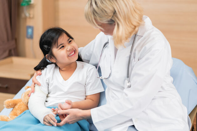 咨询小医生儿科医生和小女孩病人随身带着泰迪熊在床上病人检查疾病