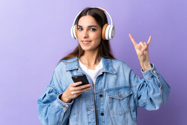科技一个年轻的白人妇女 在紫色背景下 用移动的摇滚手势听音乐手机音乐黑发