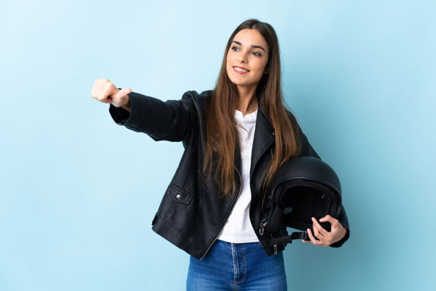 积极年轻的高加索女子手持摩托车头盔孤立在蓝色背景下竖起大拇指的手势手势好表情