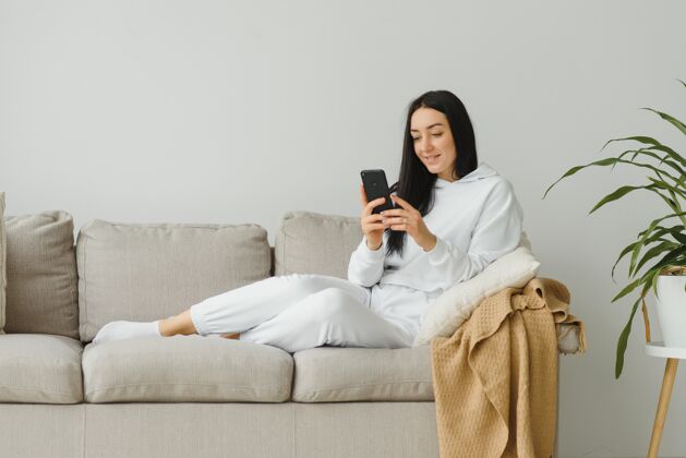 坐着一个快乐的年轻女子在家里的沙发上休息时使用手机的画像女孩快乐科技