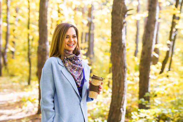享受秋天 自然 人的概念-年轻的黑发女子穿着蓝色外套站在公园里拿着杯子茶女孩学生