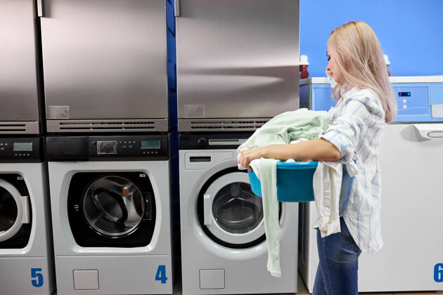 篮子一个女人独自站在带烘干机的自助洗衣房里 拿着脏衣服机器.内螺纹穿着便装的站着拿着衣服的脸盆家务职责装载