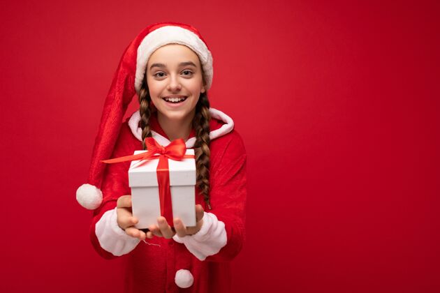庆祝照片中 美丽的快乐微笑的黑发女孩被隔离在红色背景墙上 穿着圣诞老人套装 手持红色丝带的白色礼盒 看着她照相机.副本空间黑发礼物愿望