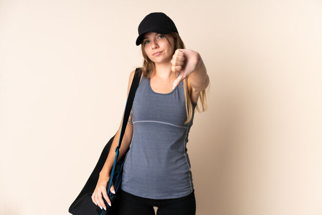拇指年轻的立陶宛运动妇女拿着一个运动包 背景是米色的 拇指朝下 表情消极年轻健身女孩