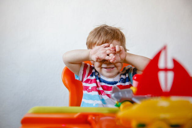 房间家里一个金发小男孩坐在一张橘黄色的儿童桌旁 桌子上放着塑料玩具 他用手捂住脸把手举起来高质量的照片游戏闭上眼睛幼儿园
