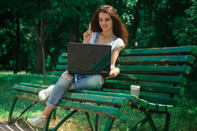 肖像可爱的微笑女士坐在公园的长椅上 看着笔记本电脑阳光桌子自然
