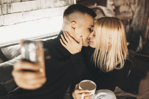 摄影师一对迷人的年轻夫妇亲吻和微笑的侧视图 而男子坐在家里的床上用智能手机自拍一起情侣女朋友