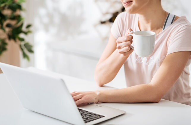 室内明亮的新鲜高兴坐在桌边喝着咖啡 用笔记本电脑浏览着一些食谱 这是一位放松的美丽女士科技早餐阅读