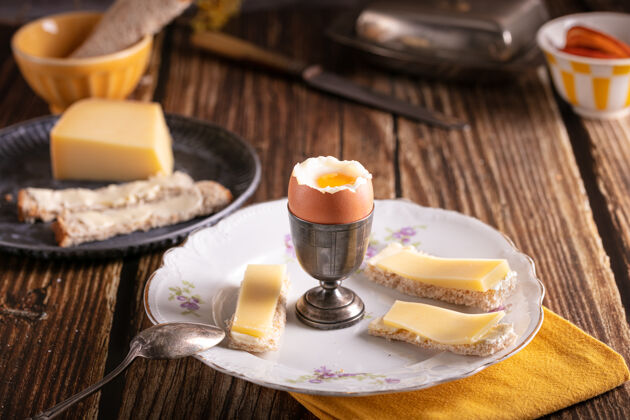 面包木桌上放着煮鸡蛋的银蛋杯 还有面包和奶酪烹饪早餐木头