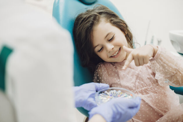 小可爱的小女孩微笑着帮助儿童口腔医生选择她未来的牙齿手术器械手套护理办公室