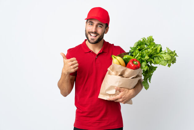 外卖一个留着胡子的年轻人拿着装满蔬菜的袋子站在白色的地板上竖起大拇指积极手势营养