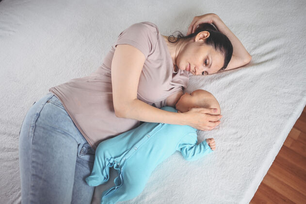 家庭年轻的母亲躺在床上 抱着一个穿着蓝色连体衣的可爱的新生婴儿 用母乳给他喂奶乳房食物可爱