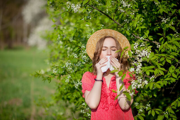 成人年轻女孩在盛开的树前用纸巾吹鼻子打喷嚏花粉打喷嚏吹