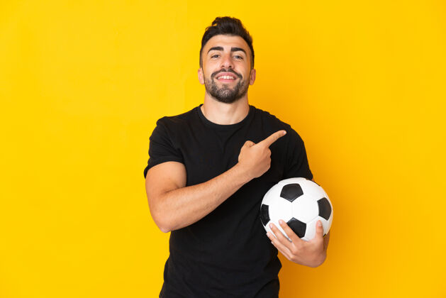 展示高加索男子在孤立的黄色背景下用足球指着侧面运动员指点多彩