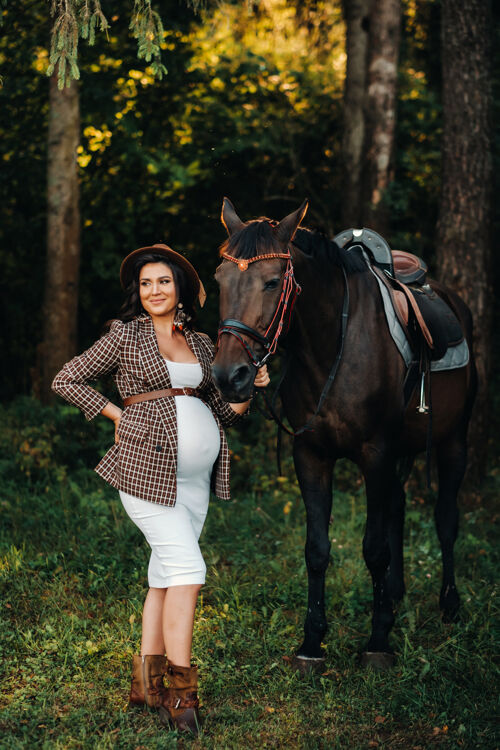怀孕的妈妈一个大肚子的怀孕女孩戴着帽子在森林里的马旁边自然时尚穿白色衣服和棕色夹克的女孩妈妈女性森林
