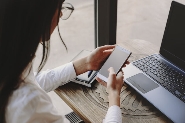 专业女性的手在使用智能手机进行互联网交易和坐在咖啡馆桌子旁的笔记本电脑女性创意笔记本电脑