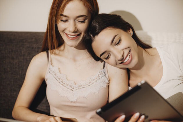 肩膀一位留着黑色长发的美丽年轻女子的画像 一边笑着一边看着她的平板电脑屏幕 一边把头靠在她红发的女性朋友身上在线姐姐朋友