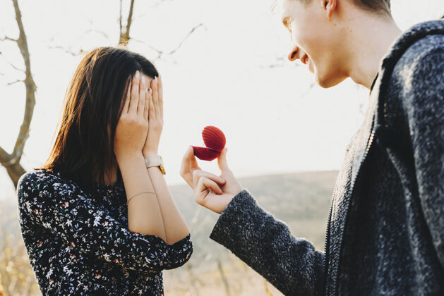 女人侧视图英俊的年轻男子微笑着拿着小戒指盒 同时用手捂着脸向匿名女子求婚快乐礼物订婚
