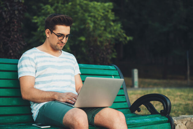 自由空间一位年轻英俊的自由职业者坐在公园的海滩上 一边用笔记本电脑工作无线眼镜户外