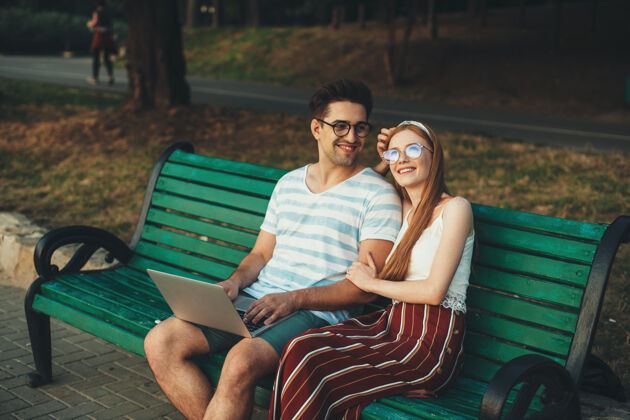 成人可爱的年轻夫妇坐在长凳上笑着 而红发女孩在户外男人年轻男朋友