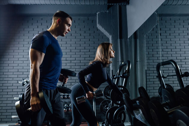运动员在黑暗的现代健身房锻炼时 穿着运动服的强壮的男男女女用哑铃做运动的侧视图物理水平努力