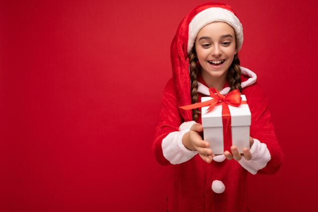 惊喜美丽快乐微笑的黑发女孩隔着红墙 穿着圣诞老人的行头 手里拿着一个白色的红包礼盒功能区空空间愿望圣诞青少年