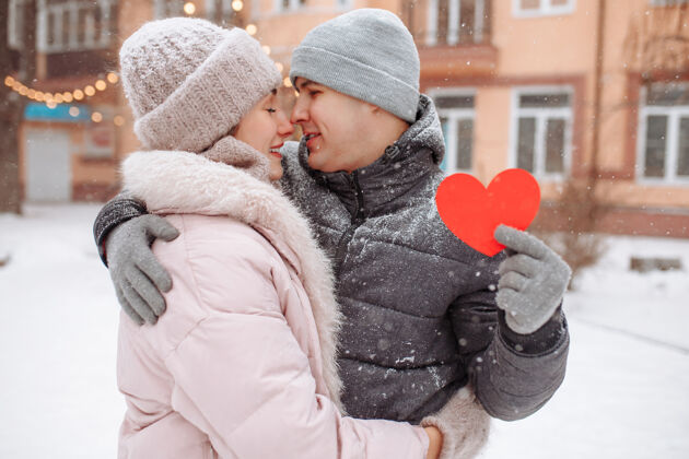 男性情人节的概念 相爱的情侣在冬天的雪地里亲吻拥抱帕克·杨男人拿着一个红色的纸心和他的女朋友一起庆祝情人节一对情侣在一起感觉很温暖举行在一起女性