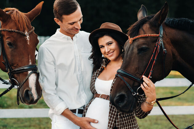 父母一个戴帽子的怀孕女孩和一个穿白衣服的男人站在白色栅栏旁的马旁边马怀孕女人