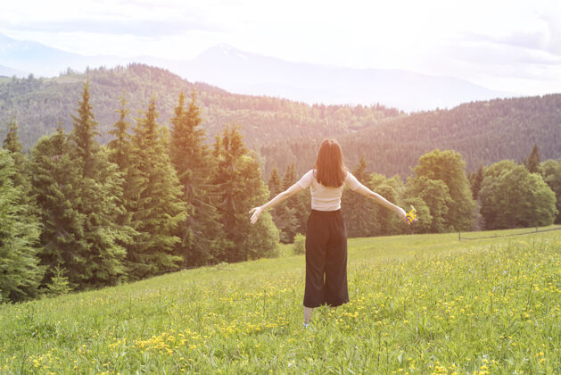 女人一个年轻的女人 手里拿着一束花 张开双臂站在花园里草地森林和山脉背景背面查看生活方式宽阔快乐