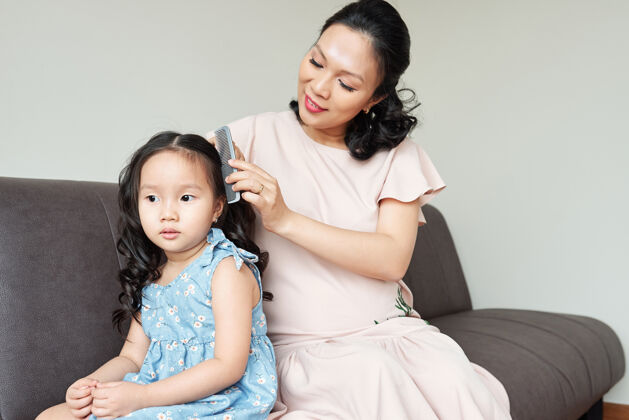 亚洲人母亲梳卷发时 坐在沙发上沉思的小女孩坐着家庭爱