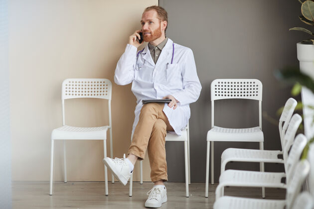 使用电话穿白大褂的男医生在医院休息时坐在椅子上讲手机 用平板电脑室内商务互联网