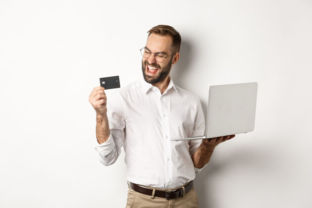 Laptop在线购物满意吗帅哥看信用卡下单后上网 用笔记本电脑 站在白色背景上男代理正式