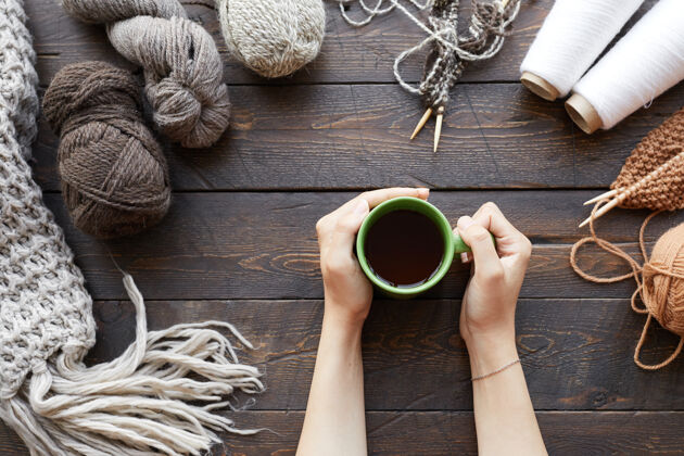 饮料特写镜头：一个女人坐在木桌旁 手里拿着毛线球和成品羊毛衣服 喝着热茶纱线冬天放松