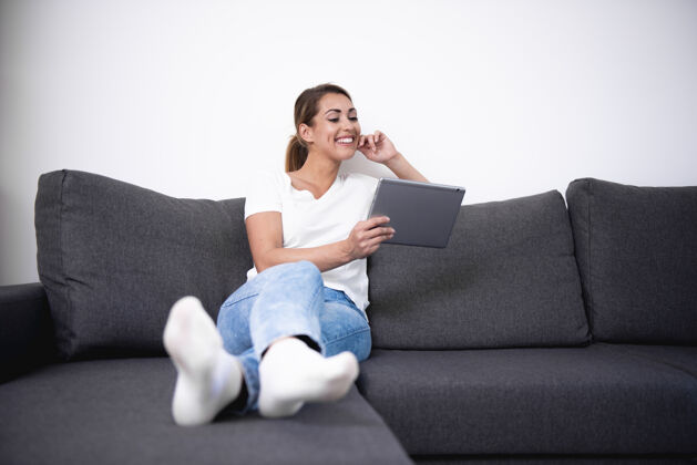 放松美丽的女人在客厅的沙发上用她的平板电脑网络美丽谈话