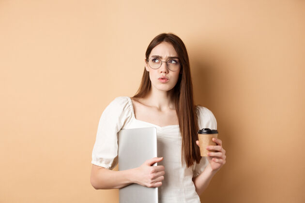 眼镜困惑而沉思的女孩拿着笔记本电脑 一边看着可疑的东西 一边喝着外卖咖啡 一边站在米色的墙上眼镜漂亮女性