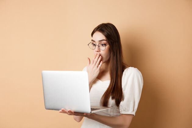 时尚震惊的女孩看着笔记本电脑屏幕 用惊讶的脸捂住嘴巴 站在米色眼镜上购物科技欢呼