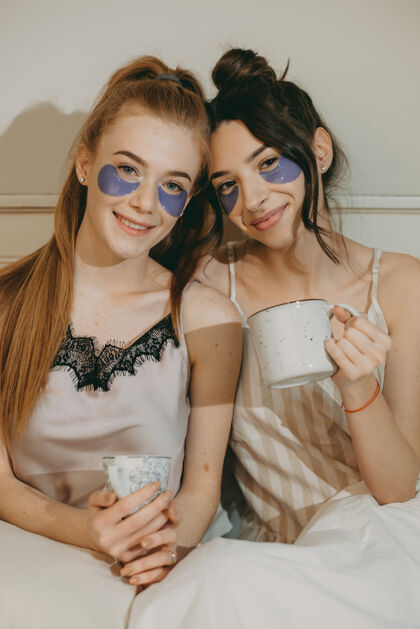 皮肤护理两个美丽的女朋友一边喝着咖啡一边放松地躺在床上 眼睛上贴着补丁年轻朋友女人