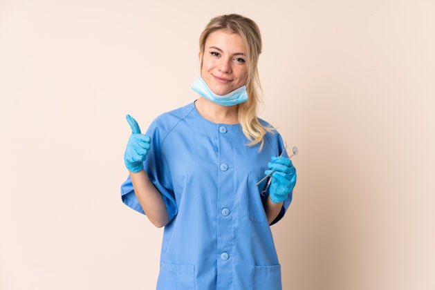 女人一个女牙医拿着工具在孤立的背景上竖起大拇指金发年轻人护理