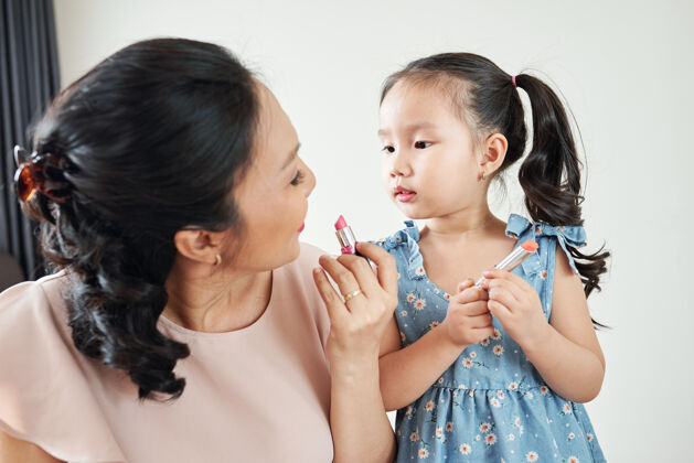 室内好奇可爱的越南小女孩看着妈妈在早晨准备好的时候涂上粉红唇膏父母女人童年