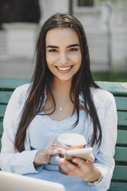 女孩照片中 一位迷人的年轻女子一边用智能手机喝咖啡 一边坐在公园的海滩上 腿上放着笔记本电脑 微笑着看着相机街道女商务人士笔记本电脑