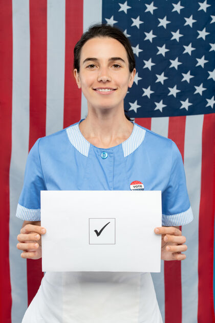 摄影棚拍摄年轻的当代微笑的女服务员 穿着蓝色制服 站在美国国旗前 向你展示她方格上打勾的选票立场美国文化职业