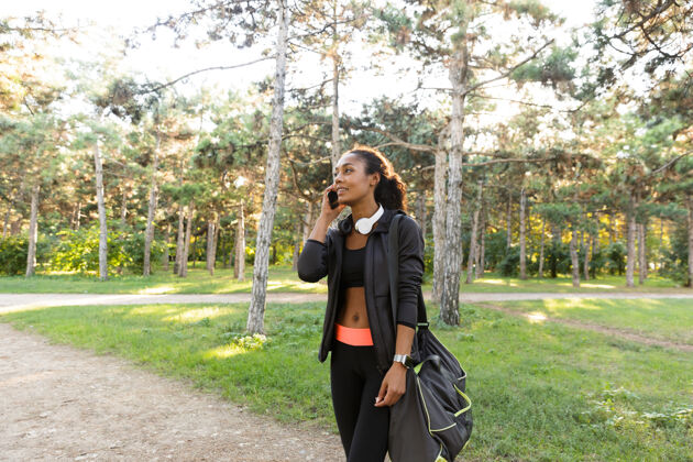 移动一个20多岁的快乐女人穿着黑色运动服 戴着耳机 一边在绿色公园散步一边用手机说话女性讲话无线