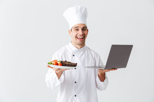 微笑兴奋的厨师穿着制服站在白色的墙上 拿着笔记本电脑 展示着一道菜菜肴帽子职业
