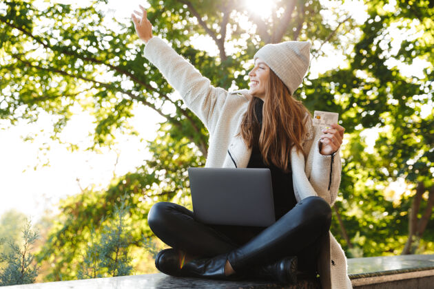 学习快乐的年轻女子穿着秋大衣 戴着秋帽坐在户外 用笔记本电脑 展示塑料信用卡使用女孩电子商务